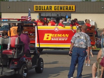 A group of people gathered around a flatbed truck decorated with banners that read 'BEMAN SHERIFF'. The truck is parked in front of a Dollar General store. Some individuals are wearing matching red shirts and appear to be part of a parade or public event. An American flag is visible on the truck, and various people in casual clothing can be seen standing or sitting nearby.