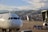 A white airplane is stationed on an airport tarmac with its nose in the foreground. Two workers dressed in safety vests are seen near baggage carts, and an aircraft boarding bridge is attached to the side. In the background, a sprawling cityscape with densely packed buildings is situated against a backdrop of mountains under a partly cloudy sky.