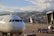 A white airplane is stationed on an airport tarmac with its nose in the foreground. Two workers dressed in safety vests are seen near baggage carts, and an aircraft boarding bridge is attached to the side. In the background, a sprawling cityscape with densely packed buildings is situated against a backdrop of mountains under a partly cloudy sky.