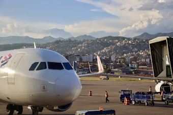 A white airplane is stationed on an airport tarmac with its nose in the foreground. Two workers dressed in safety vests are seen near baggage carts, and an aircraft boarding bridge is attached to the side. In the background, a sprawling cityscape with densely packed buildings is situated against a backdrop of mountains under a partly cloudy sky.