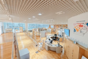 A spacious, modern library with wooden floors and shelves full of books arranged in a circular pattern. A person is seated at a computer desk in the center, with several monitors and office equipment. Large floor-to-ceiling windows allow natural light to illuminate the area. The ceiling features a grid pattern, and there are maps and informational graphics on the walls.