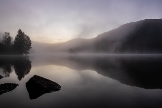 a foggy lake with trees and mountains early in the morning 