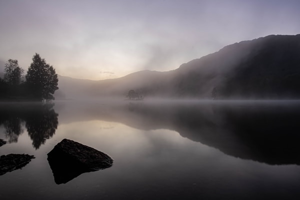 a foggy lake with trees and mountains early in the morning 
