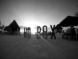 Large letters spelling out 'HO BOX' are placed prominently on a sandy beach. Thatched-roof huts and tables occupy the area with a few people present. The image appears to be taken during sunset with the sun setting on the horizon over the ocean.