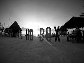 Large letters spelling out 'HO BOX' are placed prominently on a sandy beach. Thatched-roof huts and tables occupy the area with a few people present. The image appears to be taken during sunset with the sun setting on the horizon over the ocean.