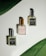 Three perfume bottles with the brand name 'AZALEEN' are arranged in a triangular pattern on a light surface. The top left bottle is labeled 'DYNAMIC' with a silver cap, the top right bottle is labeled 'MILLION SCENT' with a dark cap, and the bottom bottle is labeled 'AZALEA ROSE' with a golden cap. Large green leaves border the top left and bottom right corners, adding a natural touch.