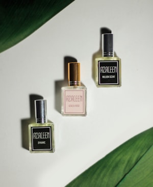 Three perfume bottles with the brand name 'AZALEEN' are arranged in a triangular pattern on a light surface. The top left bottle is labeled 'DYNAMIC' with a silver cap, the top right bottle is labeled 'MILLION SCENT' with a dark cap, and the bottom bottle is labeled 'AZALEA ROSE' with a golden cap. Large green leaves border the top left and bottom right corners, adding a natural touch.