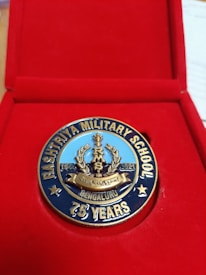 A commemorative medal celebrating the 75th anniversary of Rashtriya Military School, neatly placed inside a red presentation box. The medal features detailed engravings and inscriptions, showcasing a laurel wreath and the years '1946 - 2021'. The words 'Bengaluru' and '75 Years' are prominently displayed.