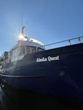 A large blue ship with the name 'Alaska Quest' displayed prominently on its side, reflecting the sunlight with clear skies in the background. The ship's windows and railings are visible, along with communication equipment on its upper structure.