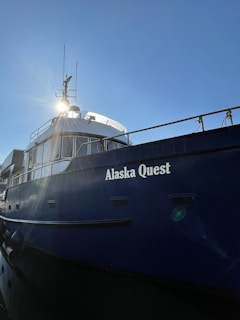 A large blue ship with the name 'Alaska Quest' displayed prominently on its side, reflecting the sunlight with clear skies in the background. The ship's windows and railings are visible, along with communication equipment on its upper structure.