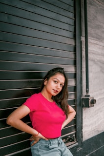 A young woman poses confidently in a vibrant pink top against a dark, industrial metal shutter. She leans slightly and gazes directly at the camera, exuding a sense of self-assurance. The background contrasts with her bright attire, highlighting her presence.