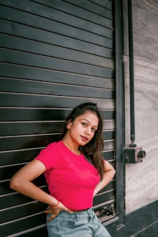 A young woman poses confidently in a vibrant pink top against a dark, industrial metal shutter. She leans slightly and gazes directly at the camera, exuding a sense of self-assurance. The background contrasts with her bright attire, highlighting her presence.
