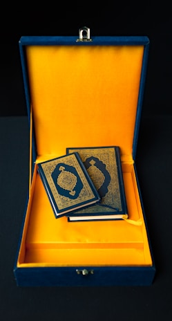 A pair of elegant books with intricate gold designs on their covers rests inside a luxurious blue and gold box. The interior of the box is lined with rich, vibrant yellow fabric, contrasting sharply with its deep blue exterior.