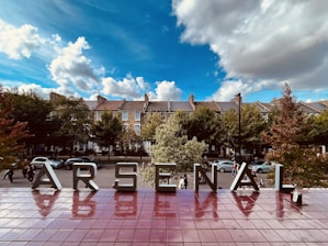 Large metal letters spelling 'Arsenal' are displayed against a backdrop of leafy trees and residential buildings under a partly cloudy blue sky. Reflections of the letters are visible on a shiny, reddish-brown tiled surface.