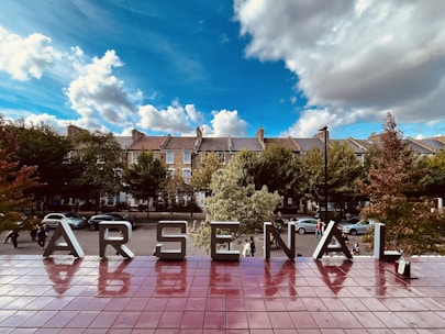 Large metal letters spelling 'Arsenal' are displayed against a backdrop of leafy trees and residential buildings under a partly cloudy blue sky. Reflections of the letters are visible on a shiny, reddish-brown tiled surface.