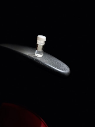 A small, transparent vial with a white cap is placed on a metallic surface against a dark background. The lighting highlights the vial and part of the metallic surface, creating a focused and dramatic contrast.