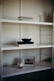 A minimalist shelf setup with a white metal frame and multiple levels. The top shelf holds a transparent box with a metal frame. The middle shelf features a white book titled 'Live Beautiful' with a dark-colored bowl placed on top. The lower shelf houses a lit candle in a glass jar and a framed picture of a solitary tree in an open field.