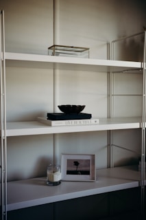 A minimalist shelf setup with a white metal frame and multiple levels. The top shelf holds a transparent box with a metal frame. The middle shelf features a white book titled 'Live Beautiful' with a dark-colored bowl placed on top. The lower shelf houses a lit candle in a glass jar and a framed picture of a solitary tree in an open field.