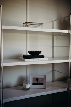 A minimalist shelf setup with a white metal frame and multiple levels. The top shelf holds a transparent box with a metal frame. The middle shelf features a white book titled 'Live Beautiful' with a dark-colored bowl placed on top. The lower shelf houses a lit candle in a glass jar and a framed picture of a solitary tree in an open field.
