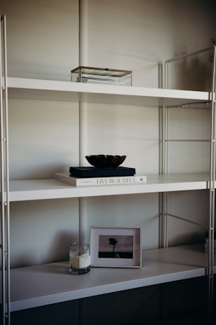 A minimalist shelf setup with a white metal frame and multiple levels. The top shelf holds a transparent box with a metal frame. The middle shelf features a white book titled 'Live Beautiful' with a dark-colored bowl placed on top. The lower shelf houses a lit candle in a glass jar and a framed picture of a solitary tree in an open field.