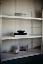 A minimalist shelf setup with a white metal frame and multiple levels. The top shelf holds a transparent box with a metal frame. The middle shelf features a white book titled 'Live Beautiful' with a dark-colored bowl placed on top. The lower shelf houses a lit candle in a glass jar and a framed picture of a solitary tree in an open field.