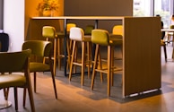 A modern, stylish cafe setting with wooden high tables and chairs in various shades of light brown and green. A vase with flowers is placed on the table, adding a touch of nature to the contemporary design. Soft natural light filters in through large windows, creating a calm and inviting atmosphere.