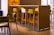 A modern, stylish cafe setting with wooden high tables and chairs in various shades of light brown and green. A vase with flowers is placed on the table, adding a touch of nature to the contemporary design. Soft natural light filters in through large windows, creating a calm and inviting atmosphere.