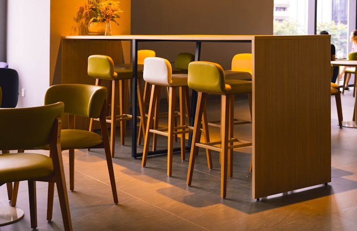 A modern, stylish cafe setting with wooden high tables and chairs in various shades of light brown and green. A vase with flowers is placed on the table, adding a touch of nature to the contemporary design. Soft natural light filters in through large windows, creating a calm and inviting atmosphere.