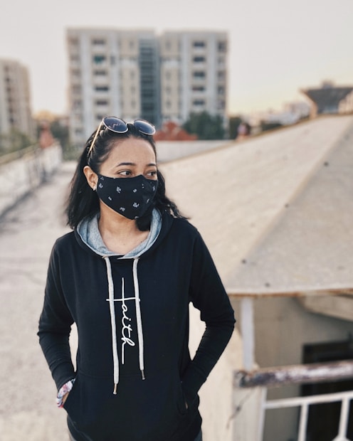 A person wearing a black hoodie with the word 'Faith' on it stands on a rooftop. They are also wearing a black mask with white patterns and sunglasses on top of their head. In the background, there are tall residential buildings, and the sun is setting or rising, casting a warm light.