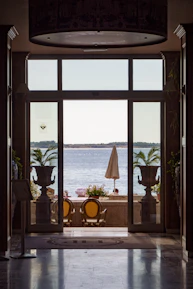 a patio with a table and chairs and a large window with a view of the ocean