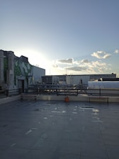 An urban rooftop setting with tiled flooring and a view of the city skyline. There are benches along the perimeter, a few scattered puddles on the ground, and a single potted plant in a terracotta pot. In the background, industrial pipes and buildings are visible under a clear sky with scattered clouds. The sun is low on the horizon, creating a soft glow.