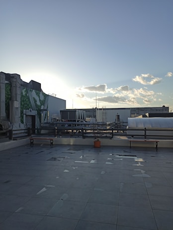 An urban rooftop setting with tiled flooring and a view of the city skyline. There are benches along the perimeter, a few scattered puddles on the ground, and a single potted plant in a terracotta pot. In the background, industrial pipes and buildings are visible under a clear sky with scattered clouds. The sun is low on the horizon, creating a soft glow.