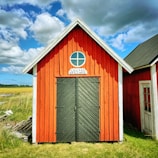 A small red wooden building with a gabled roof features a black double door and a circular window above it. The sign above the door reads 'Lillväg Skillinge'. Surrounded by grassy fields under a partly cloudy blue sky.