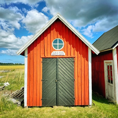 A small red wooden building with a gabled roof features a black double door and a circular window above it. The sign above the door reads 'Lillväg Skillinge'. Surrounded by grassy fields under a partly cloudy blue sky.