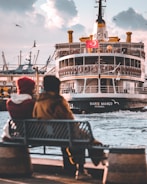 A couple sits on a bench by the waterfront, overlooking a large passenger ferry with a Turkish flag. The ferry, named 'Barış Manço', is approaching the dock, and several seagulls fly around it. The sky is partly cloudy, contributing to a serene, late afternoon atmosphere.