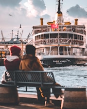 A couple sits on a bench by the waterfront, overlooking a large passenger ferry with a Turkish flag. The ferry, named 'Barış Manço', is approaching the dock, and several seagulls fly around it. The sky is partly cloudy, contributing to a serene, late afternoon atmosphere.