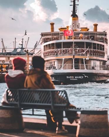 A couple sits on a bench by the waterfront, overlooking a large passenger ferry with a Turkish flag. The ferry, named 'Barış Manço', is approaching the dock, and several seagulls fly around it. The sky is partly cloudy, contributing to a serene, late afternoon atmosphere.