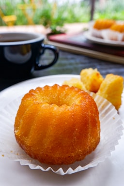 A small, golden-brown bundt cake rests in a white paper cup on a plate. Accompanying the cake are small pastry pieces, all set against a backdrop featuring a black coffee cup filled with coffee and blurred greenery in the distance.