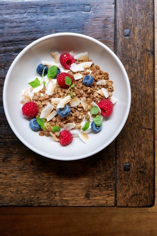 A white bowl filled with granola, raspberries, blueberries, coconut flakes, and garnished with fresh mint leaves, placed on a rustic wooden table.