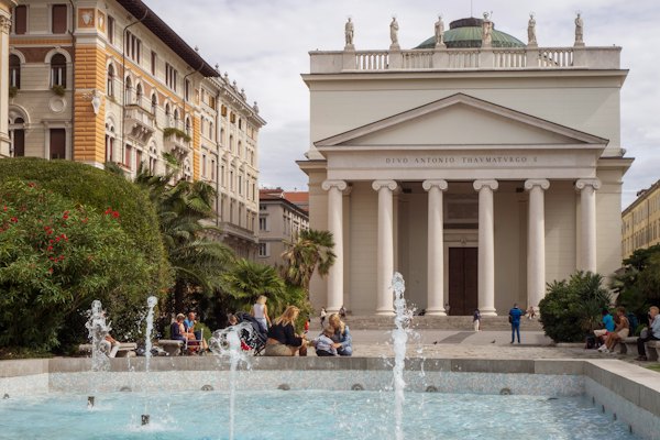 A group of people sitting around a fountain in front of a building — Trieste, Italy