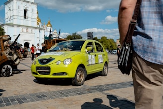 A bright green car with visible damage is parked on a paved area, with debris on its hood. Several people are gathered nearby, and in the background, a white building with gold domes is visible. The sky is partly cloudy, and a person in the foreground is holding a bag.