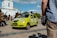 A bright green car with visible damage is parked on a paved area, with debris on its hood. Several people are gathered nearby, and in the background, a white building with gold domes is visible. The sky is partly cloudy, and a person in the foreground is holding a bag.
