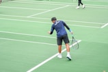 A person wearing a blue shirt and black shorts is holding a tennis racket on a green tennis court. The person appears to be preparing for a serve or adjusting their position. Another player is visible in the background, wearing black pants and preparing a swing with their racket.