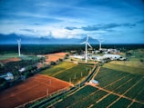 A scenic landscape featuring expansive agricultural fields with neatly arranged crops in various shades of green and brown. Tall wind turbines stand prominently, harnessing wind energy against a backdrop of distant mountains under a vast, cloud-covered sky. A small cluster of buildings is nestled among the fields, adding a sense of habitation to the otherwise natural scene.