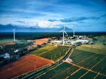 A scenic landscape featuring expansive agricultural fields with neatly arranged crops in various shades of green and brown. Tall wind turbines stand prominently, harnessing wind energy against a backdrop of distant mountains under a vast, cloud-covered sky. A small cluster of buildings is nestled among the fields, adding a sense of habitation to the otherwise natural scene.