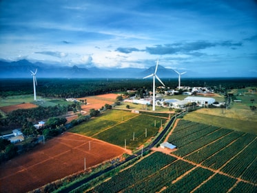 A scenic landscape featuring expansive agricultural fields with neatly arranged crops in various shades of green and brown. Tall wind turbines stand prominently, harnessing wind energy against a backdrop of distant mountains under a vast, cloud-covered sky. A small cluster of buildings is nestled among the fields, adding a sense of habitation to the otherwise natural scene.