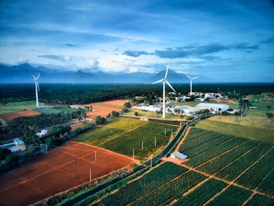 A scenic landscape featuring expansive agricultural fields with neatly arranged crops in various shades of green and brown. Tall wind turbines stand prominently, harnessing wind energy against a backdrop of distant mountains under a vast, cloud-covered sky. A small cluster of buildings is nestled among the fields, adding a sense of habitation to the otherwise natural scene.
