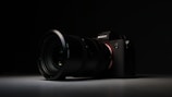 A high-quality camera with a large lens is set against a dark background. The camera is positioned at an angle, with the lens prominently in focus. The overall lighting is low, highlighting the camera's sleek design and details on the lens and body.