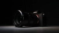 A high-quality camera with a large lens is set against a dark background. The camera is positioned at an angle, with the lens prominently in focus. The overall lighting is low, highlighting the camera's sleek design and details on the lens and body.