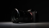 A high-quality camera with a large lens is set against a dark background. The camera is positioned at an angle, with the lens prominently in focus. The overall lighting is low, highlighting the camera's sleek design and details on the lens and body.